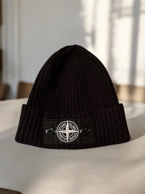 Svart ribbad mössa från Stone Island - Svart ribbstickad mössa från Stone Island med klassisk logotyp framtill på en svart patch med knappar. Tillverkad i 100% bomull och designad i Italien. Mössan har uppvikt kant och en clean, stilren look som passar till streetwear.