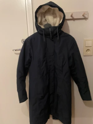Mörkblå parkas från Helly Hansen - Säljer en mörkblå parkas från Helly Hansen med huva och mysigt ljust teddyfoder. Jackan har dragkedja, snörning vid huvan och stora fickor framtill. Perfekt för kalla dagar med sitt vind- och vattenavvisande material. Lång modell som täcker bra och håller värmen.