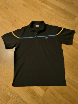 Svart piké från Lacoste med blå och gula detaljer - Svart pikétröja från Lacoste med klassisk krage och korta ärmar. Framsidan har en blå linje över bröstet och gula linjer på axlarna. Ikoniska Lacoste-loggan på vänster bröst och liten krokodiltagg i nederkant. Materialet är bomull och passformen är normal.