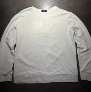 Ralph Lauren Vit Tröja - Säljer en Ralph Lauren Vit Tröja med ett snyggt USA flaggmärke med textur. Den är sjukt skön, riktigt snygg och passar till allt. Storlek: L 14/16 men kan annars användas som S. Skriv vid frågor eller kom med bud!