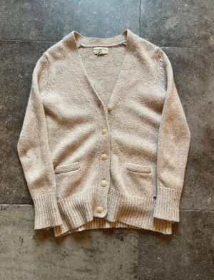 Denim supply cardigan - Säljer en beige stickad kofta från Ralph Lauren Denim & Supply. Koftan har v-ringning, ribbade muddar, fyra knappar framtill och två fickor. Perfekt för lager-på-lager och chill stil. Passar dig som gillar klassisk och enkel design.