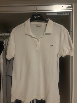 Vit pikétröja från GANT, 176 cm - Vit pikétröja från GANT med klassisk krage och korta ärmar. Tröjan har en liten broderad logga på bröstet och knappar vid halsen. Tillverkad i mjuk bomull som känns skön mot huden. Perfekt för dig som gillar stilrena och enkla plagg.