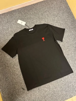 Svart Ami Paris t-shirt med rött hjärta - Svart t-shirt från Ami Paris i mjuk bomull. Klassisk rund hals och korta ärmar. På bröstet finns ett broderat rött hjärta med bokstaven A. Enkel och stilren design som ger en cool vibe till din outfit.