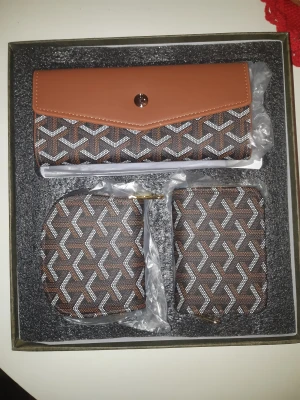 Brunt och mönstrat plånboksset - Snyggt set med plånbok och två mindre fodral i brunt och svart med grafiskt mönster. Plånboken har ett lock med knapp och de mindre fodralen har dragkedja. Perfekt för att hålla koll på kort, mynt och småsaker. Materialet är syntet och designen är riktigt modern.