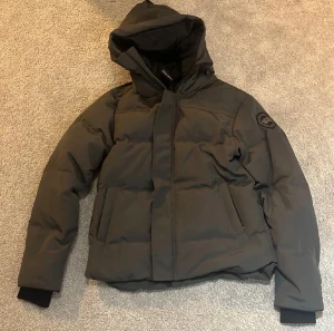 Grå pufferjacka från Canada Goose - Säljer en grå pufferjacka från Canada Goose med huva och svart patch på ärmen. Jackan har dragkedja och knappar framtill, två stora fickor och ribbade muddar vid ärmslut. Perfekt för kalla dagar och har en clean, modern look.