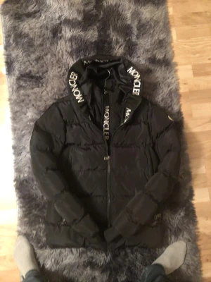 Svart Moncler pufferjacka med huva - Svart pufferjacka från Moncler i storlek S med stor huva och tydligt Moncler-tryck på huvkanten. Jackan har dragkedja framtill, två sidofickor och klassisk Moncler-logga på ärmen. Insidan har en cool comic strip-etikett. Perfekt för kalla dagar.