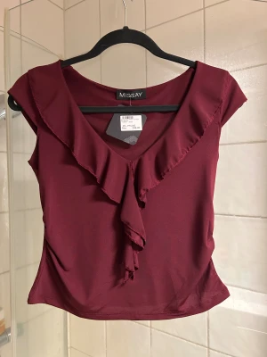 Vinröd volangblus från Missäy - Supersnygg vinröd blus från Missay med volangdetaljer längs v-ringningen och framtill. Modellen är croppad och har korta ärmar. Materialet är mjukt och följsamt, troligen polyester. Perfekt att styla med jeans eller kjol för en trendig look.