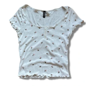 Vit blommig Topp - Supersöt vit topp från hm med små rosa blommor och gröna blad. Den har korta ärmar, v-ringad hals med spetskant och dekorativa knappar framtill. Materialet är mjukt och ribbat, vilket gör den extra bekväm och feminin. Perfekt för dig som gillar romantisk och girly stil.