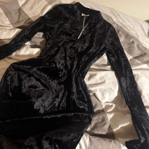 Svart sammetsklänning med dragkedja - Supersnygg svart sammetsklänning med lång ärm och tight passform. Klänningen är kort och har en cool silverfärgad dragkedja framtill med ringdetalj. Perfekt för dig som vill sticka ut med en stilren men ändå trendig look. XS