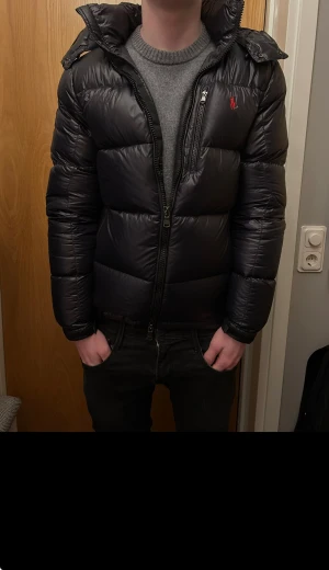 Svart pufferjacka från Polo Ralph Lauren - Snygg svart pufferjacka från Polo Ralph Lauren med röd logga på bröstet. Jackan har huva, flera dragkedjor och en quiltad design som ger en cool och modern look. Perfekt för kalla dagar och har en normal passform. Nästan helt ny, knappt använd. Säljer då den är för liten för mig.