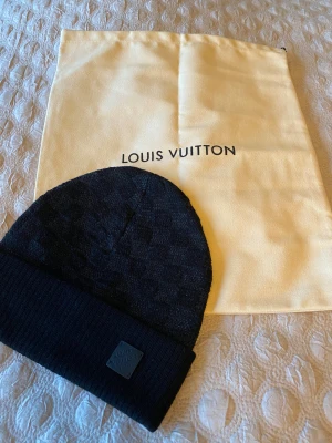 Svart Louis Vuitton mössa  - Snygg svart mössa från Louis Vuitton med diskret rutigt mönster och ribbad uppvikt kant. Märkesplakett framtill och exklusiv känsla. Materialet är mjuk ullblandning som håller dig varm och stilren under kalla dagar.