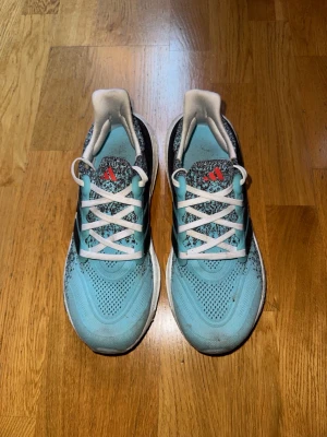 Adidas Ultraboost sneakers, stl 41 1/3 - Snygga Adidas Ultraboost sneakers i ljusblått med svarta och vita detaljer samt röd logga på plösen. Ovandelen är stickad med mönstrad design och har klassiska tre svarta ränder på sidan. Skorna har vit och svart platt sula och vita snören för en fräsch look. Storlek 41 1/3. Sporadiskt använda och endast lite smutsiga, inga skador. 