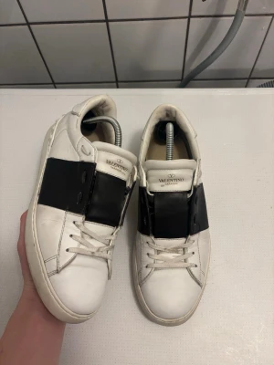 Valentino Garavani Open sneakers - Säljer ett par snygga Valentino Garavani Open sneakers i vitt skinn med bred svart läderdetalj över mitten. Klassisk rund tå, platt sula och vita snören. Märkeslogga på plösen och stilren design som sticker ut. Storlek 42 i bra kvalité