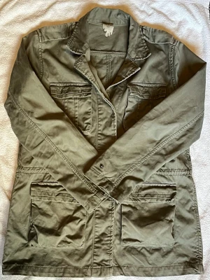 Monki Field jacket - As fet Field jacket ifrån Monki, med flera stora fickor framtill och klassisk krage. Jackan har snörning i midjan för att justera passformen och stängs med knappar