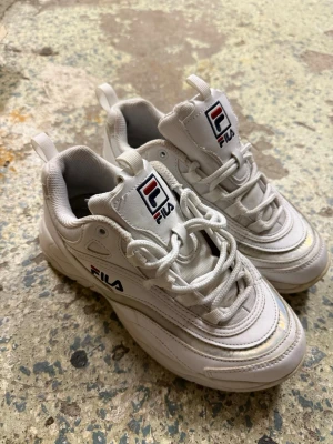 Vita chunky sneakers från FILA - Säljer ett par vita chunky sneakers från FILA i storlek 38. Skorna har en cool, vågig sula och detaljer i syntetmaterial med textilfoder. FILA-logga på sidan och plösen, samt snörning framtill. Perfekt för dig som gillar sportig och trendig stil.