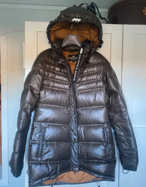 Brun pufferjacka Mountain Works M - Säljer en brun, glansig pufferjacka från Mountain Works, modell Foxy Lady 75. Jackan har huva, dragkedja och stora ringar på dragkedjorna. Fodret är brunt och jackan är quiltad med långa ärmar. Perfekt för kalla dagar.