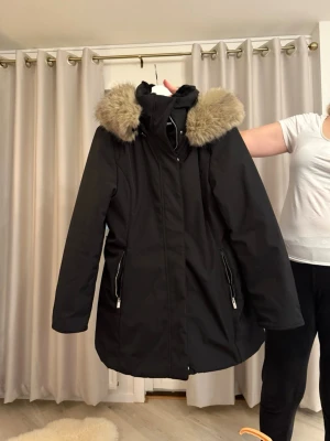 Svart parkas med fuskpäls - Svart parkas med huva och fluffig beige fuskpäls runt kragen. Jackan har långa ärmar, två fickor med dragkedja och en stilren, något figursydd form. Perfekt för kalla dagar med sin varma och snygga look.