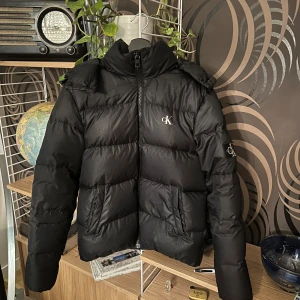 jacka från Calvin Klein  - Svart pufferjacka från Calvin Klein med huva och dragkedja framtill. Jackan har CK-logga på bröstet och ärmen, samt två stora fickor. Perfekt för kalla dagar med sin tjocka vaddering och stilrena look.