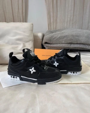 Louis Vuitton svarta sneakers - Svarta Louis Vuitton sneakers med vita detaljer och LV-logga på hälen. Snyggt mönstrad mesh och skinn på ovandelen, samt blommig detalj på sidan. Platt sula i svart och vitt, snörning och rund tå. Perfekt för dig som gillar exklusiv streetstyle.