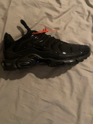 Nike Air Max Plus svarta sneakers - Nike Air Max Plus sneakers i helsvart med Tn Air-detalj på sulan och röd tag. Snygg vågig design på ovandelen, snörning och platt sula. Materialet är syntet och mesh för skön komfort. Perfekt för dig som gillar streetwear och sportig stil.