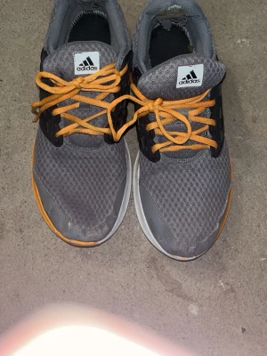 Grå Adidas sneakers med orange detaljer - Säljer ett par grå Adidas sneakers med mesh-ovandel, svarta plastdetaljer och orange snörning. Skorna har vit och orange sula samt svart hälparti. Perfekta för dig som gillar sportig stil och vill sticka ut med färgglada detaljer.