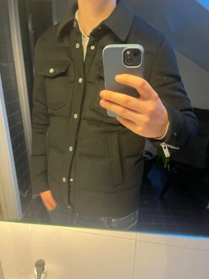 Dunjacka, Kashmir/Ull från Moose Knuckles - Svart dunjacka med yttertyg i kashmir och ull från Moose Knuckles med klassisk krage, tryckknappar och två bröstfickor med lock. Jackan har quiltad design och patch med logga på ärmen. Perfekt för kalla dagar och har en snygg, stilren look. Galje kommer med, kvitto finns. Använd enbart ett fåtal gånger.