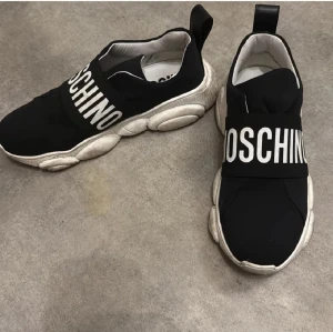 Svarta sneakers från Moschino - Säljer ett par svarta sneakers från Moschino med bred vit logga över foten. Skorna har chunky vit sula med unik bubblig design och är tillverkade i syntetmaterial. Slip-on modell med dragflik bak för enkel påtagning. Riktigt statement-piece för dig som gillar streetstyle. Billigare vid upphämtning!!!