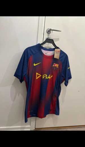 Ingen sko på bilden - Bilderna visar en FC Barcelona matchtröja från Nike, inte skor. Tröjan har röd och blå randigt mönster, gul Nike-logga och klubbmärke på bröstet. Materialet är lätt och ventilerande polyester, perfekt för fotboll.