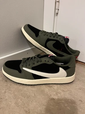 Travis Scott Skor  - Nike Air Jordan 1 Low Travis Scott i olivgrönt och svart med vit omvänd swoosh. Skorna har röda broderade detaljer på hälen och klassisk snörning. Ovandelen är i skinn och mocka, med en platt sula i beige. Perfekt för dig som vill sticka ut med en ikonisk sneaker.