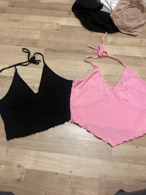 2 croppade linne-toppar, svart & rosa - Två croppade linne-toppar med smala axelband och v-ringning. En svart topp med broderat blommönster och en rosa ribbad topp med spetsig nederkant. Båda har knyt i nacken och är perfekta för sommaren. Den svarta har en skada som visas på bilden 