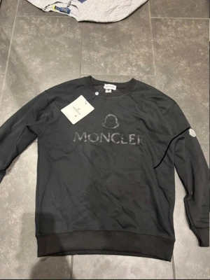 Svart sweatshirt från Moncler - Säljer en svart sweatshirt från Moncler med diskret logga och emblem på ärmen. Tröjan har rund halsringning och ribbade muddar vid ärmslut och nederkant. Perfekt för en stilren och avslappnad look.