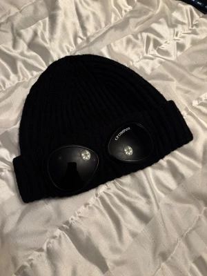 Svart C.P. Company mössa - Svart ribbstickad mössa från C.P. Company med ikoniska svarta goggle-linser framtill. Mycket bra skick !!!!!!