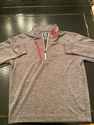 Grå half zip tröja från FootJoy - Säljer en gråmelerad tröja från FootJoy med athletic fit. Tröjan har en vit half zip framtill och en röd dragkedja på bröstet. FJ-logga i nacken och på kragen. Materialet är mjukt och syntetiskt, perfekt för golf.