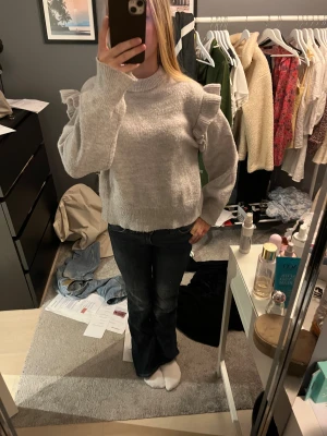 Grå stickad tröja med volangärmar - Superfin ljusgrå stickad tröja med volangdetaljer på axlarna och långa ärmar. Tröjan har en rund halsringning och ribbade muddar vid ärmslut och nederkant. Perfekt för dig som gillar en söt och trendig look.