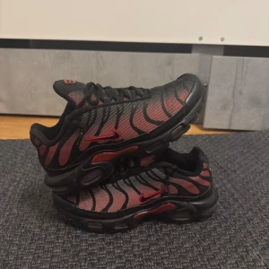 Nike Air Max Plus svarta/röda sneakers - Nike Air Max Plus/Tn sneakers med svart och röd mesh-överdel, svarta vågformade detaljer och röd Swoosh på sidan. Platt sula med synliga Air-enheter och röda inslag på undersidan.  Pris kan diskuteras