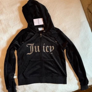 Juicy Couture svart hoodie, storlek L ny med prislapp - Svart hoodie med dragkedja framtill. Hoodien har texten "Juicy" i strass. Luvan har dragsko.  Storlek: L Skick: Helt ny med prislapp (1100kr)