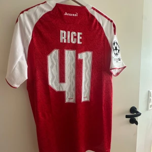 Arsenal Rice Hemmatröja 25/26 - Säljer en officiell Arsenal fotbollströja från Adidas med Rice 41 på ryggen. Tröjan är röd med vita ärmar, har Champions League-märke på ärmen och detaljer i guld. Materialet är lätt och andas, perfekt för match eller träning. Snyggt mönster i tyget och klassisk Arsenal-logga på bröstet.