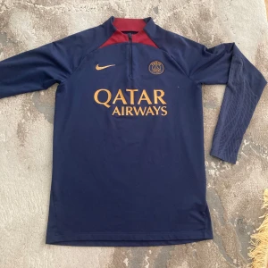 PSG långärmad fotbollströja Nike S - Paris Saint-Germain långärmad fotbollströja från Nike i mörkblått med röda detaljer vid halsen. Tryckt Qatar Airways-logga i guld på bröstet och PSG-emblem i guld. Materialet är lätt och andas, perfekt för träning eller match.