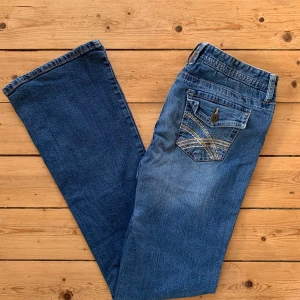 Blå bootcut jeans från rue21 W32 L32 - Snygga blå jeans från rue21 med bootcut passform och klassisk femficksdesign. Bakfickorna har dekorativa sömmar och knappdetalj. Jeansen har dragkedja och knapp framtill samt coolt mönstrat innerfoder i midjan. Perfekta för en avslappnad och trendig look.    Storlek W32 L32