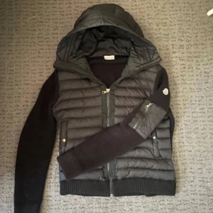 Moncler Cardigan - Svart cardigan från Moncler med stickade ärmar och ribbade muddar. den har quiltad front, huva och dragkedja samt Moncler-logga på ärmen. Materialet är en mix av dun och stickad polyester, perfekt för kyliga dagar. Snygg och sportig look med praktiska fickor.