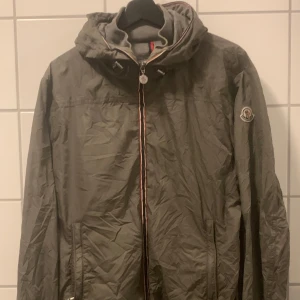 Grå vindjacka från Moncler med huva - Snygg grå vindjacka från Moncler med huva och dragkedja framtill. Jackan har Moncler-logga på ärmen och kan packas ihop till en smidig väska med röd, vit och blå axelrem. Perfekt för dig som vill ha en lätt och stilren jacka med sportig känsla.     (UK	US	Moncler size 38	M	2 40	L	3 42	XL	4 44	XXL	5)