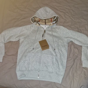 Grå hoodie från Burberry med zip - Snygg ljusgrå hoodie från Burberry med klassiskt rutigt foder i huvan. Tröjan har hel dragkedja, snörning i huvan och två stora fickor framtill. Jag säljer den pågrund av att den är lite för liten för mig och den är inte köpt i Sverige så kunde tyvärr inte lämna tillbaka den 