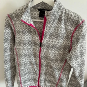 Fleecetröja från Everest storlek 146/152 - Mysig gråvit fleecejacka från Everest med nordiskt mönster och rosa dragkedja. Jackan har lång ärm och hög krage, perfekt för kyliga dagar.Storlek 146/152 och i fint skick.