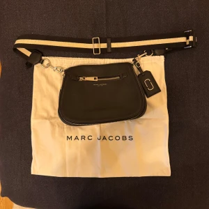 Svart axelväska från Marc Jacobs - Snygg svart axelväska från Marc Jacobs i äkta skinn med justerbar randig axelrem. Väskan har silverfärgade dragkedjor, ett ytterfack med dragkedja och flera innerfack. Perfekt för dig som vill ha en stilren och praktisk väska med exklusiv känsla. Använd 2 gånger så väldigt bra skick, som ny.