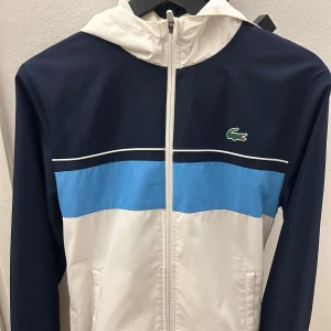  vindjacka från Lacoste  - Snygg vindjacka från Lacoste Sport i blått, vitt och ljusblått med dragkedja och huva. Jackan har klassisk krokodillogga på bröstet och sportig design med färgblock. Perfekt för dig som gillar streetwear och sportig stil.