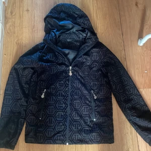 Svart vindjacka från Philipp Plein  - Svart vindjacka från Philipp Plein med hel dragkedja, huva och två fickor med dragkedja. Jackan har ett mönster med Philipp Plein-loggor över hela ytan och en glansig finish. Perfekt för dig som gillar streetstyle och exklusiva detaljer.