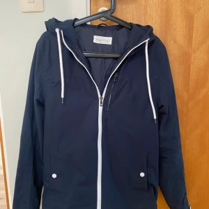 Mörkblå Windbreaker - En riktigt snygg och bekväm mörkblå Windbreaker från Pier One med dragkedja och huva. Med snygga vita detaljer och en inneficka. Passar mig inte längre men har knappt används. Bara fråga om du undrar något, priset kan diskuteras 