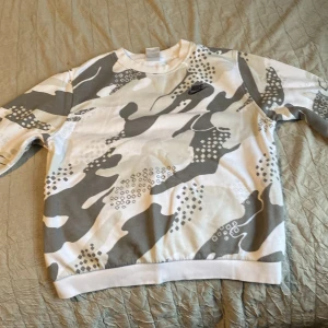 Nike sweatshirt med grått camomönster XL - Nike sweatshirt i storlek XL med grått och vitt camouflagemönster. Tröjan har rund halsringning, långa ärmar och Nike-logga broderad på bröstet. Perfekt för dig som gillar streetwear och vill sticka ut med en unik look.