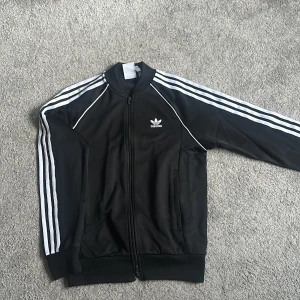 Svart Adidas track kofta med vita ränder - Klassisk svart Adidas track jacket med vita ränder längs ärmarna och broderad logga på bröstet. Jackan har dragkedja framtill, ribbade muddar och två fickor. Tillverkad i polyester för en sportig och clean look.