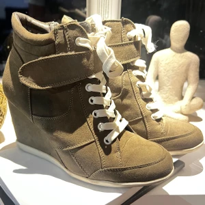 Khakifärgade sneakers med kilklack och snörning - Isabel marant liknande skor. Snygga khakifärgade sneakers med kilklack och vita snören. Skorna har en cool look med rund tå, mockaliknande material och kardborreband upptill. Perfekta för dig som vill ha lite extra höjd men ändå en avslappnad stil.